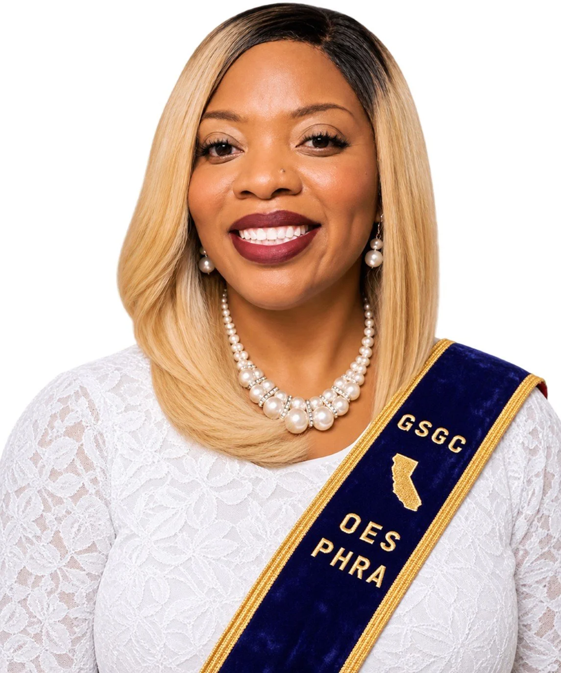 An older woman wearing a white lace dress with pearls and a navy sash with gold trim, standing against a colorful abstract background.