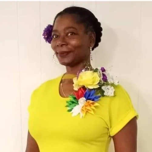 A woman wearing a bright yellow shirt with a large floral decoration on her shoulder, smiling with a purple flower in her hair and earrings, standing against a white wall.