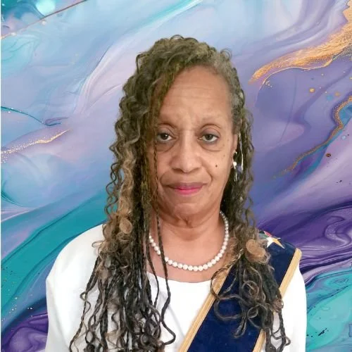 A woman with long, curly gray hair, wearing a white top, pearl necklace, and a dark blue sash with a golden stripe, standing in front of a colorful, abstract background.