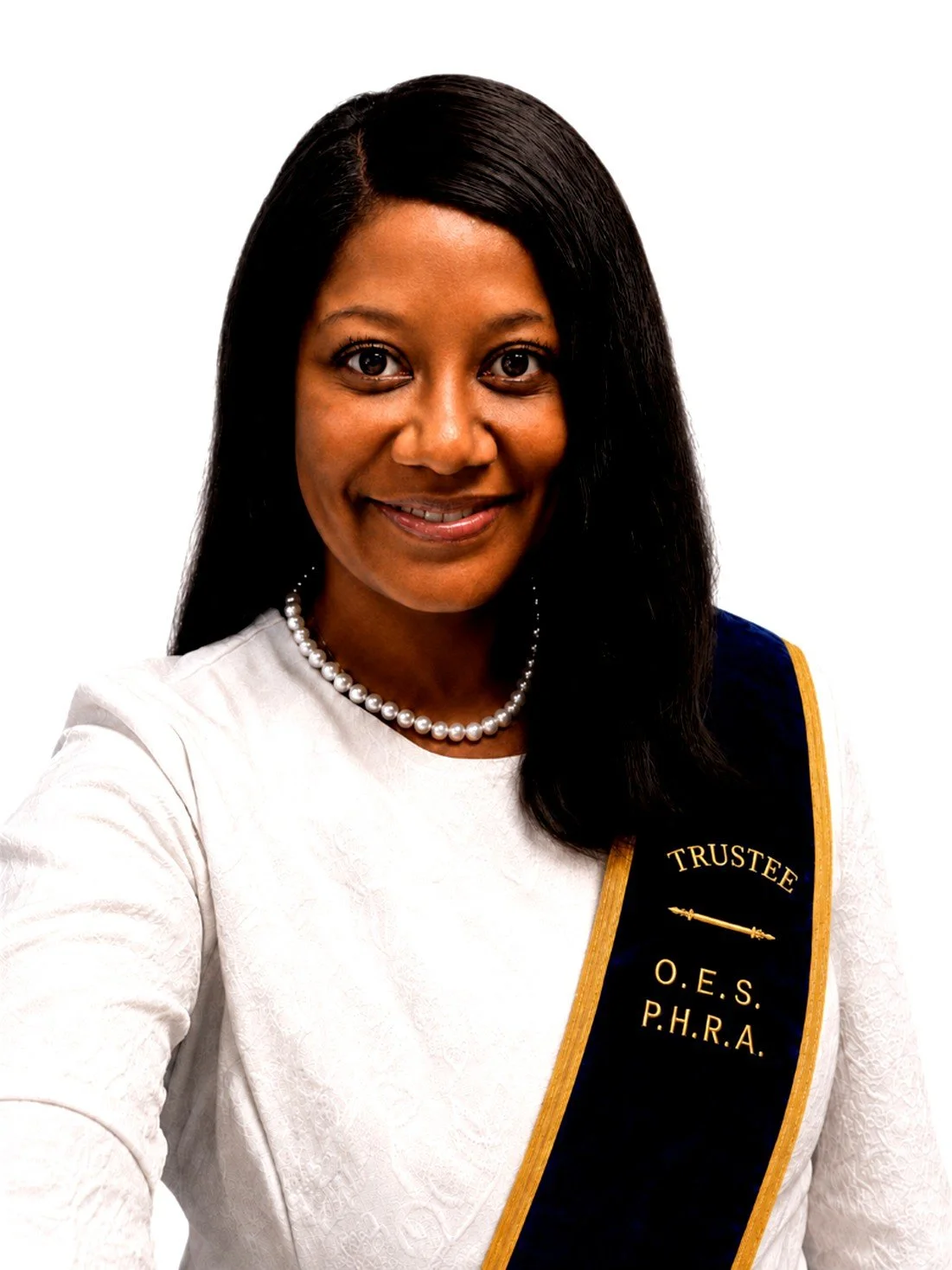 A woman with long, curly gray hair, wearing a white top, pearl necklace, and a dark blue sash with a golden stripe, standing in front of a colorful, abstract background.