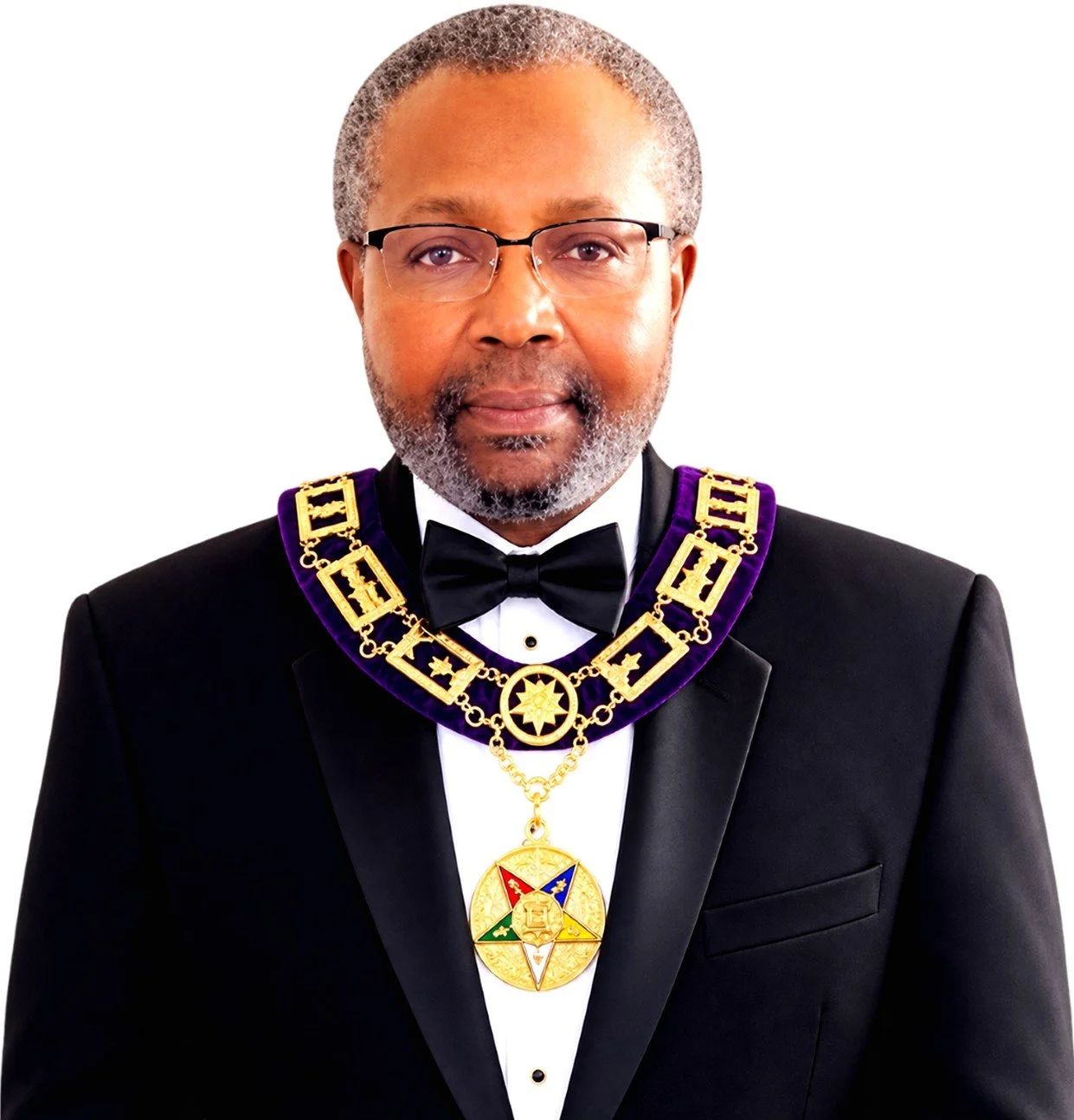 A man in formal attire wearing glasses and a ceremonial chain with a star pendant, standing against an abstract blue and purple background.