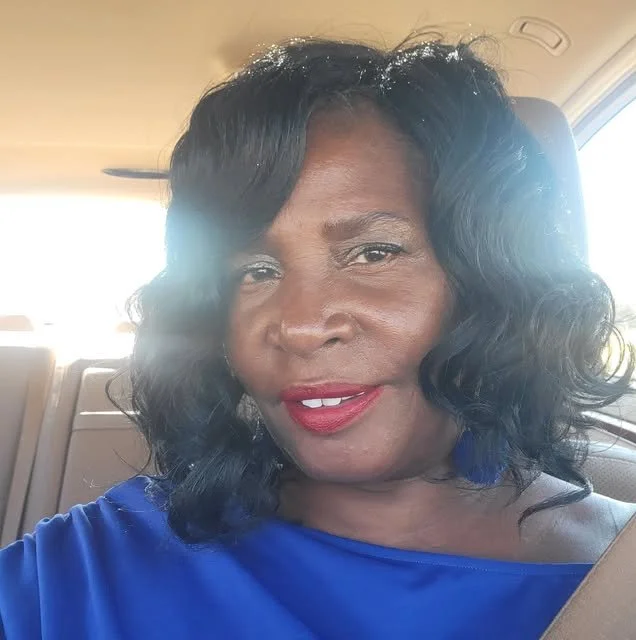 A woman with dark, wavy hair and red lipstick taking a selfie inside a car, wearing a blue top.