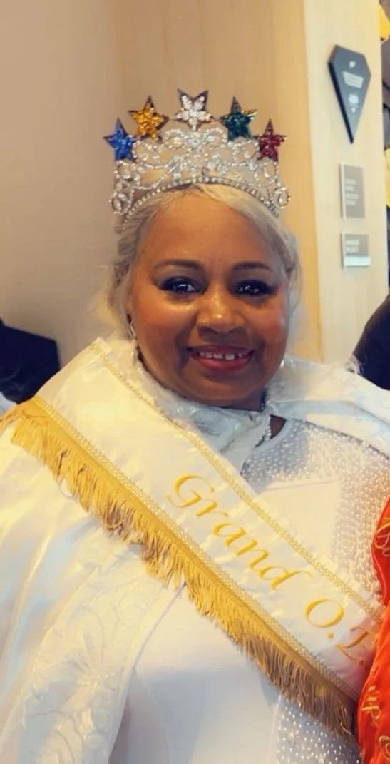 A woman wearing a crown decorated with multicolored stars, a white sash with gold trim that reads 'Grand Queen,' and a white dress with intricate detailing.