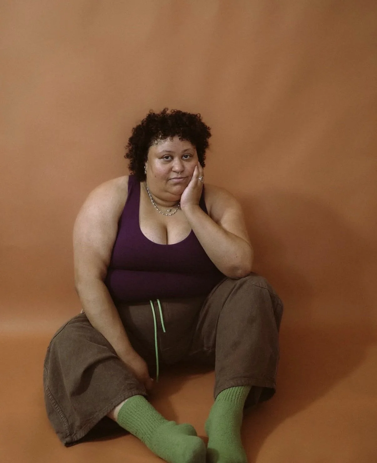 Photo shows Mandy, a woman sitting in front of a terracotta colored background. She has green plants and socks on, as well as an earth toned purple shirt. She is the holistic skincare specialist and esthetician behind faceplant.