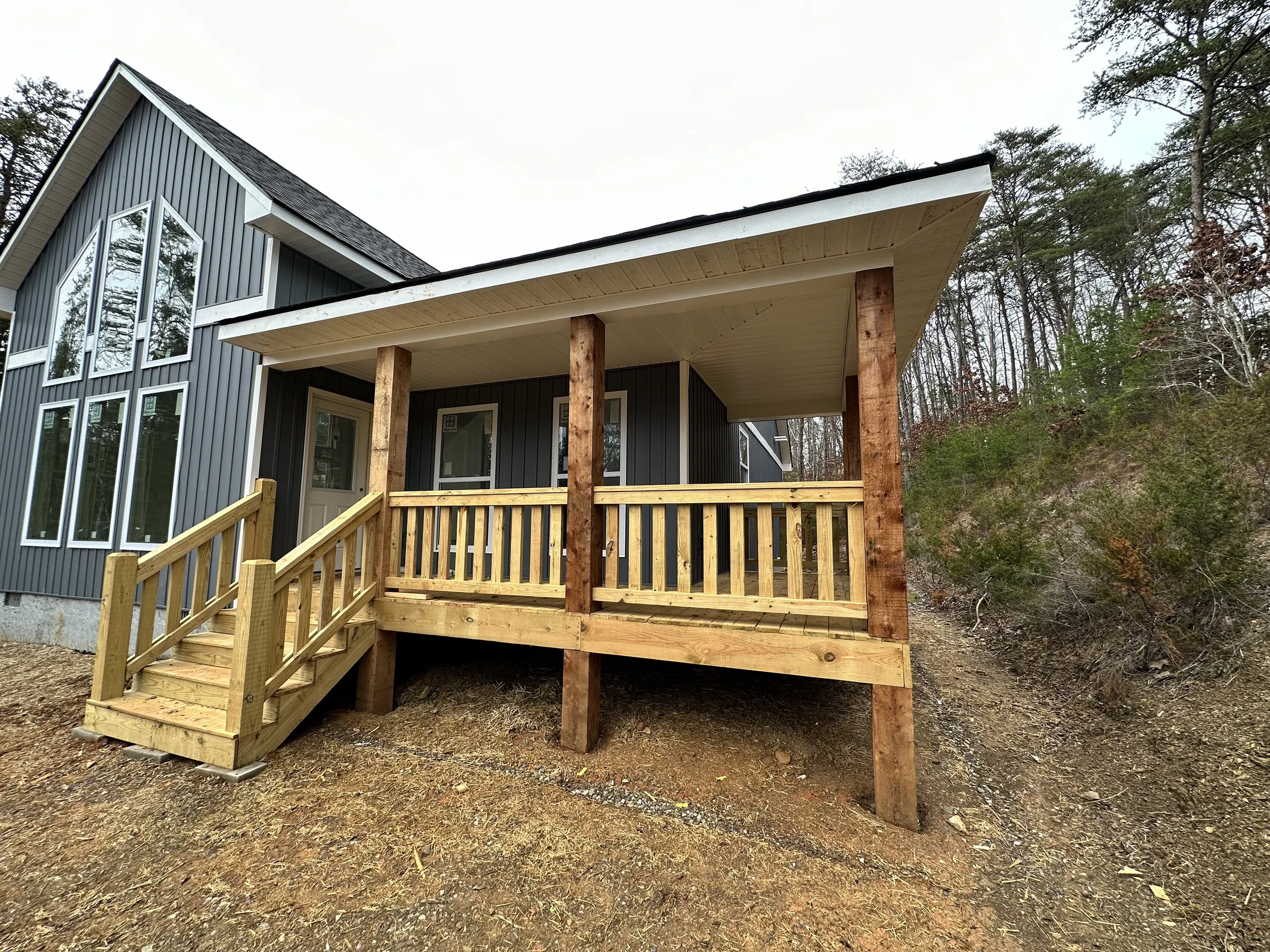 Wooden deck with outdoor dining table and chairs, a gas grill, and a blue house with a door, surrounded by tall green trees.