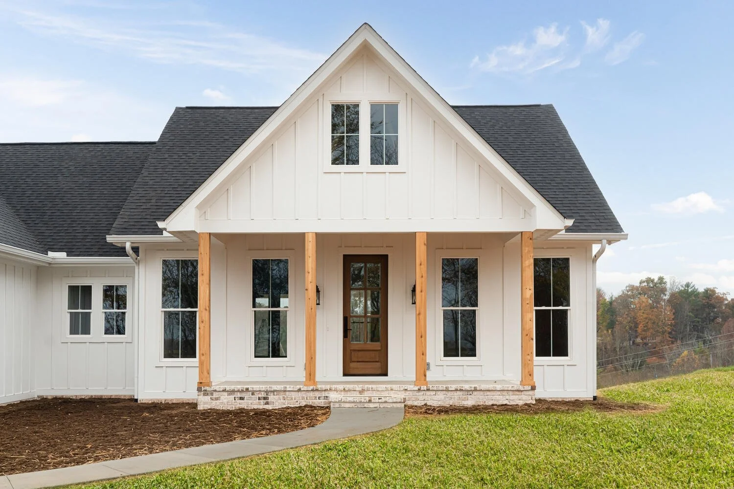 A house under construction with a blue crane lifting roofing materials, situated on a green field with trees and hills in the background.