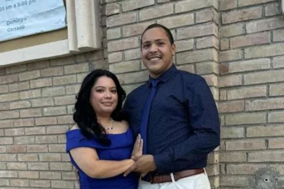 A woman and a man standing close together, smiling, against a brick wall. The woman has shoulder-length black hair and is wearing a blue dress. The man has short dark hair and is wearing a dark blue shirt with a tie and white pants.