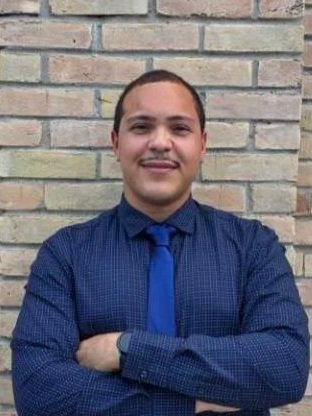 A young man with short hair, wearing a blue shirt and tie, standing in front of a brick wall.