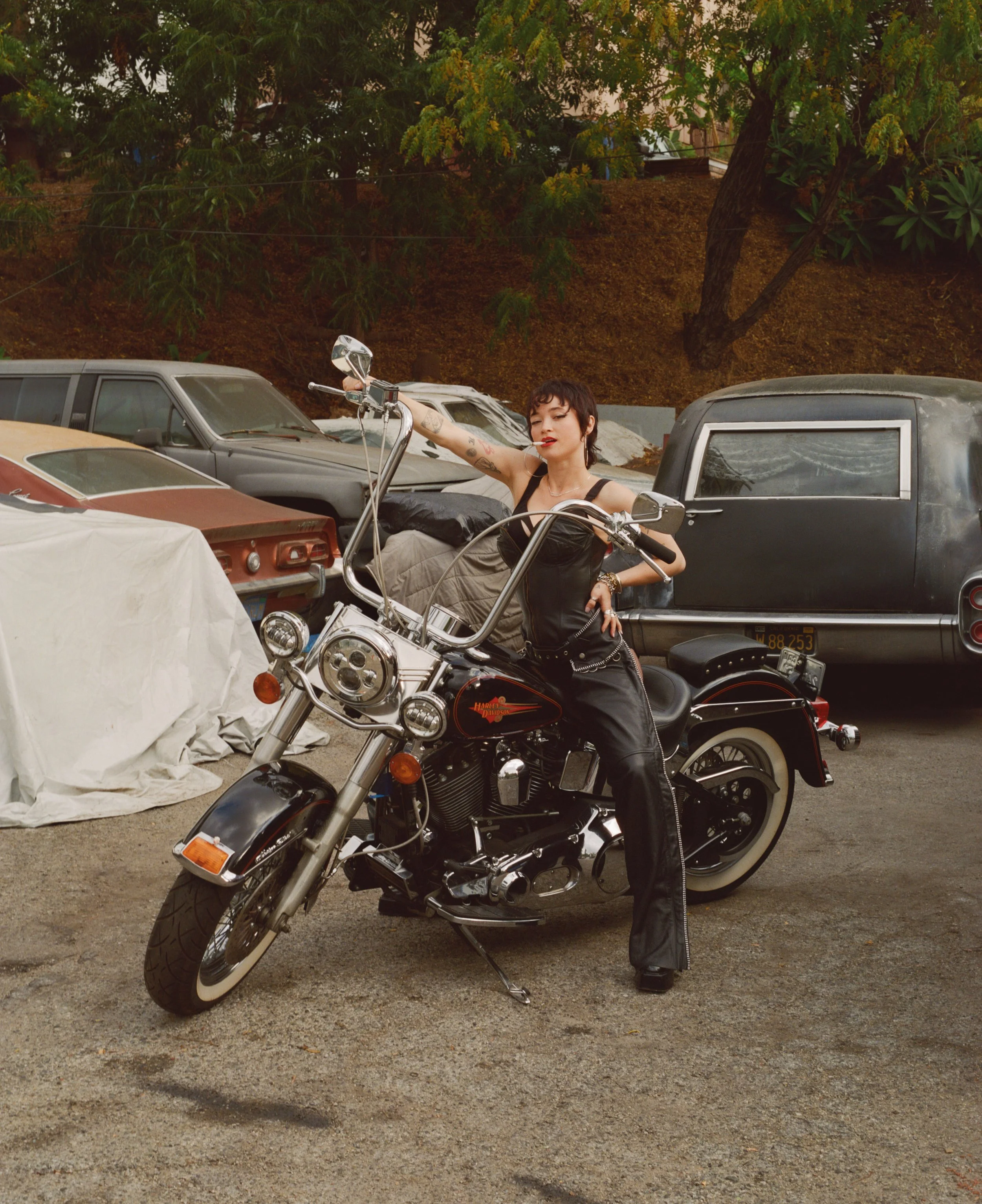 Shooting with Tyana P in Los Angeles.
A woman with short dark hair and tattoos, dressed in black leather, poses on a black Harley-Davidson motorcycle in a vintage car lot with old cars and a dirt hill with trees in the background.