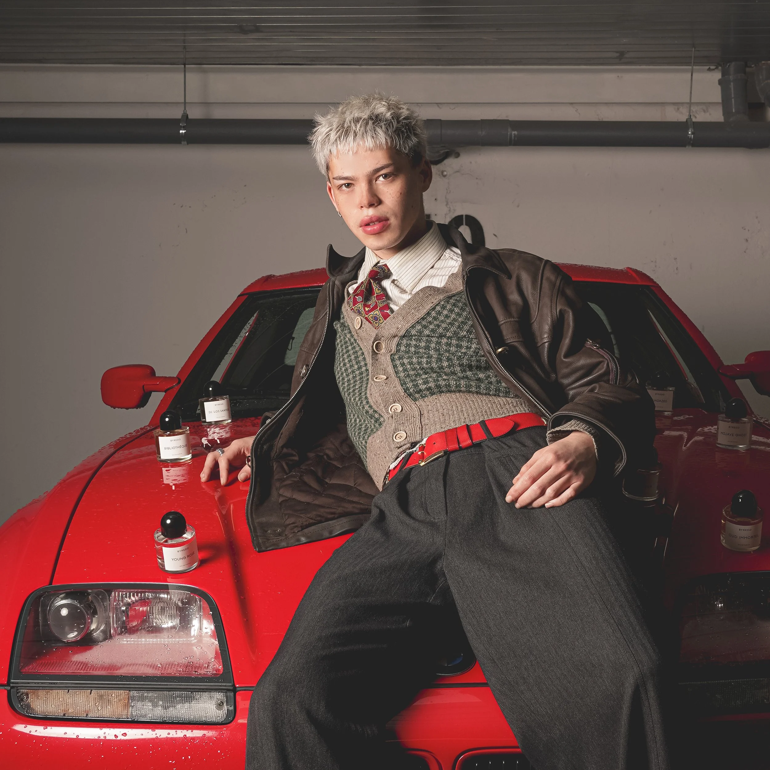 A young man with platinum blonde hair leaning against a red sports car in a parking garage. He's wearing a patterned sweater vest, a shirt, a tie, a leather jacket, and dark pants.