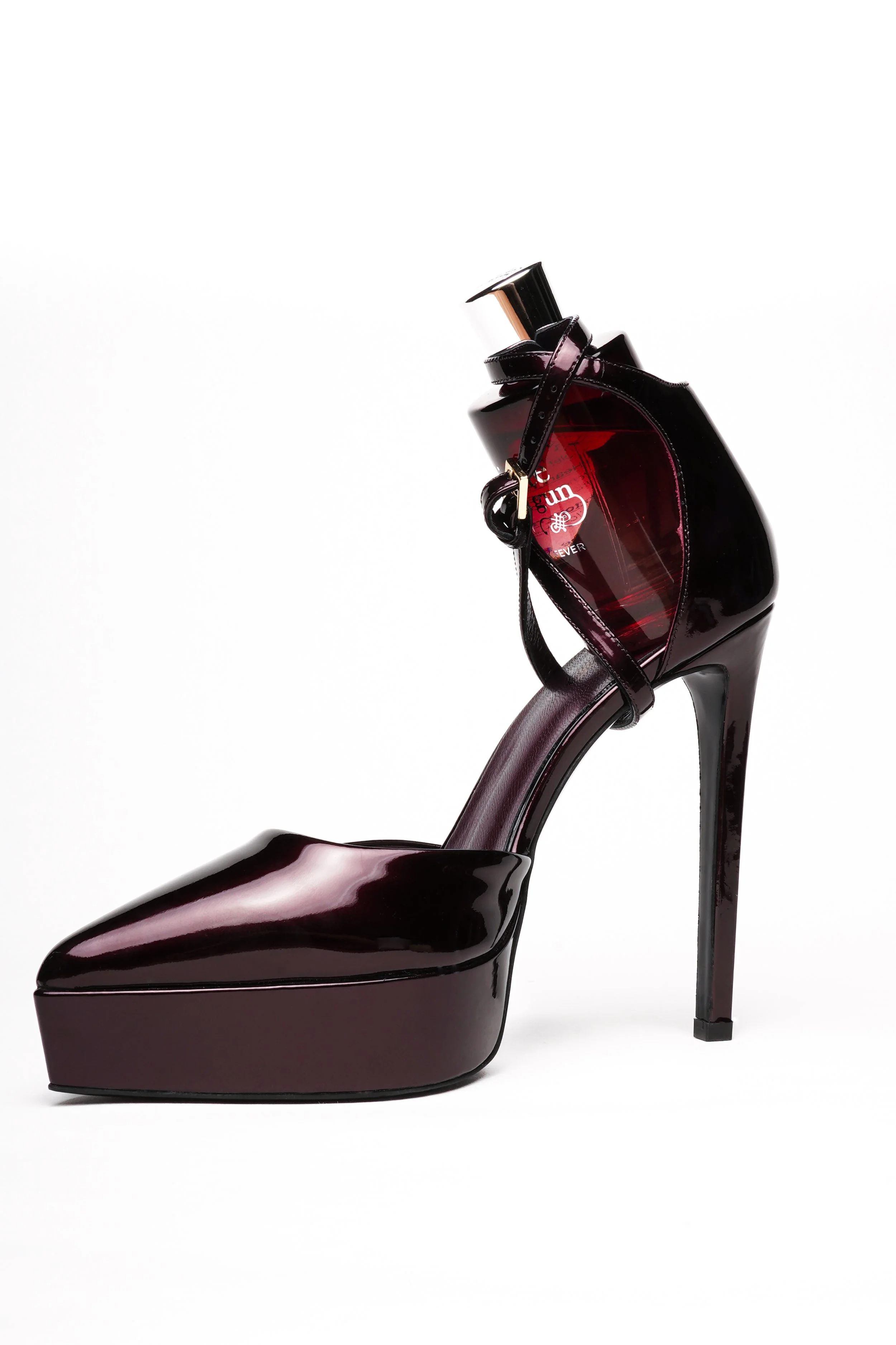 A high-heeled shoe with a platform, shiny dark burgundy color, and an ankle strap, with a perfume bottle integrated into the ankle strap.