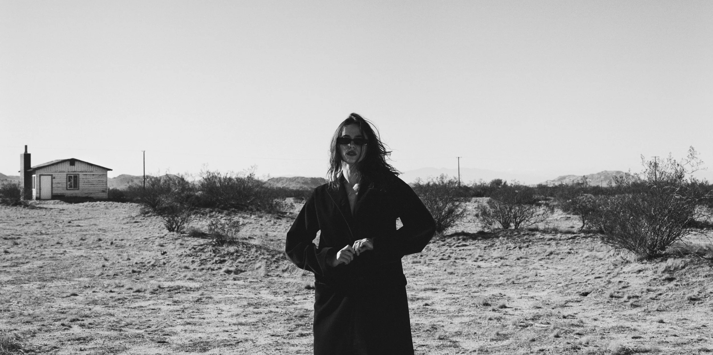Shooting with the actress Tu Morrow in the desert of Los Angeles.
A woman with wavy hair wearing sunglasses, a dark jacket, and a skirt standing in a barren desert landscape with sparse bushes and a small wooden house in the background.