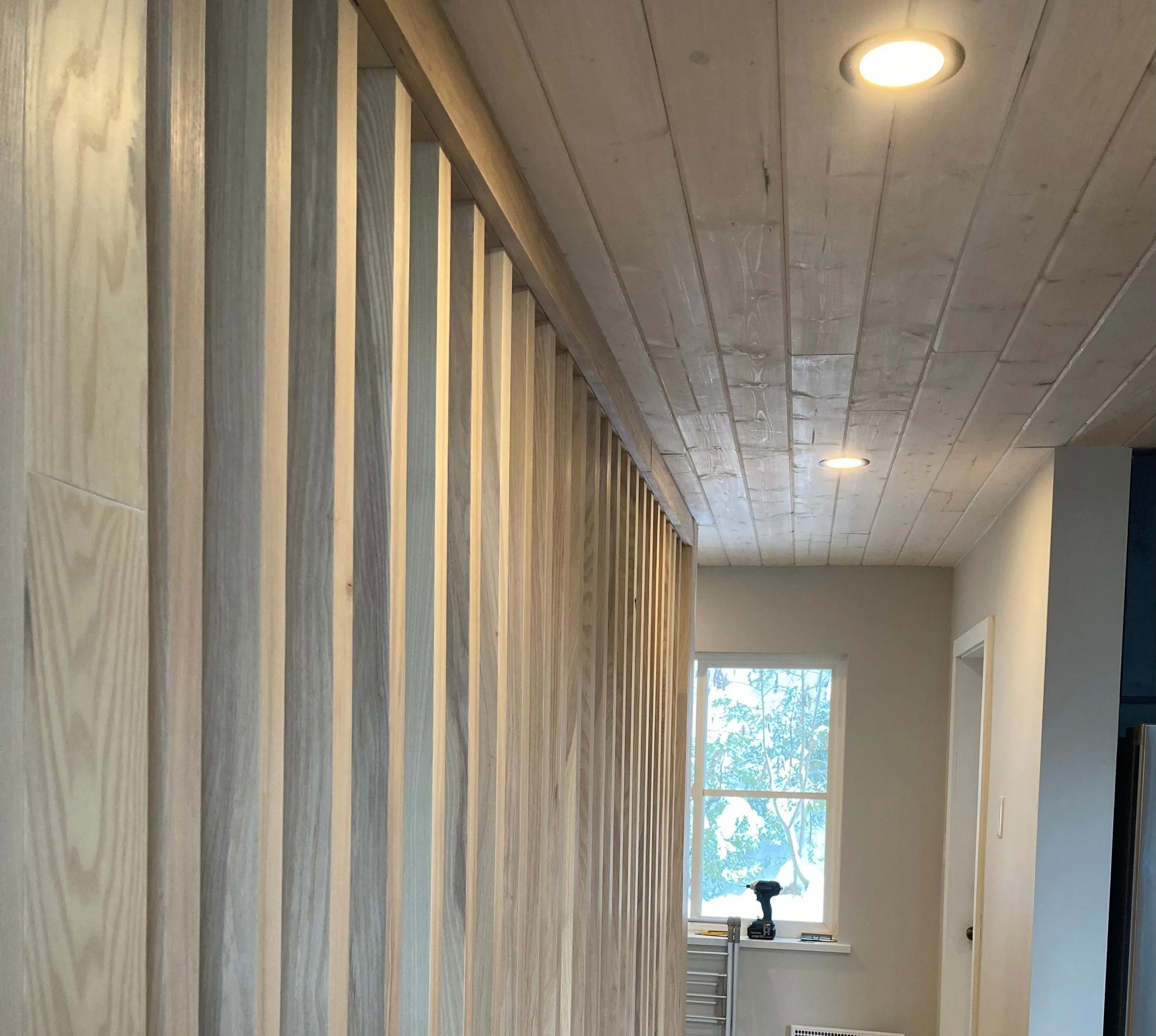 Interior of a room under construction with a vertical wooden slat wall, a window showing trees outside, and a drilled hole on the ceiling with recessed lighting.