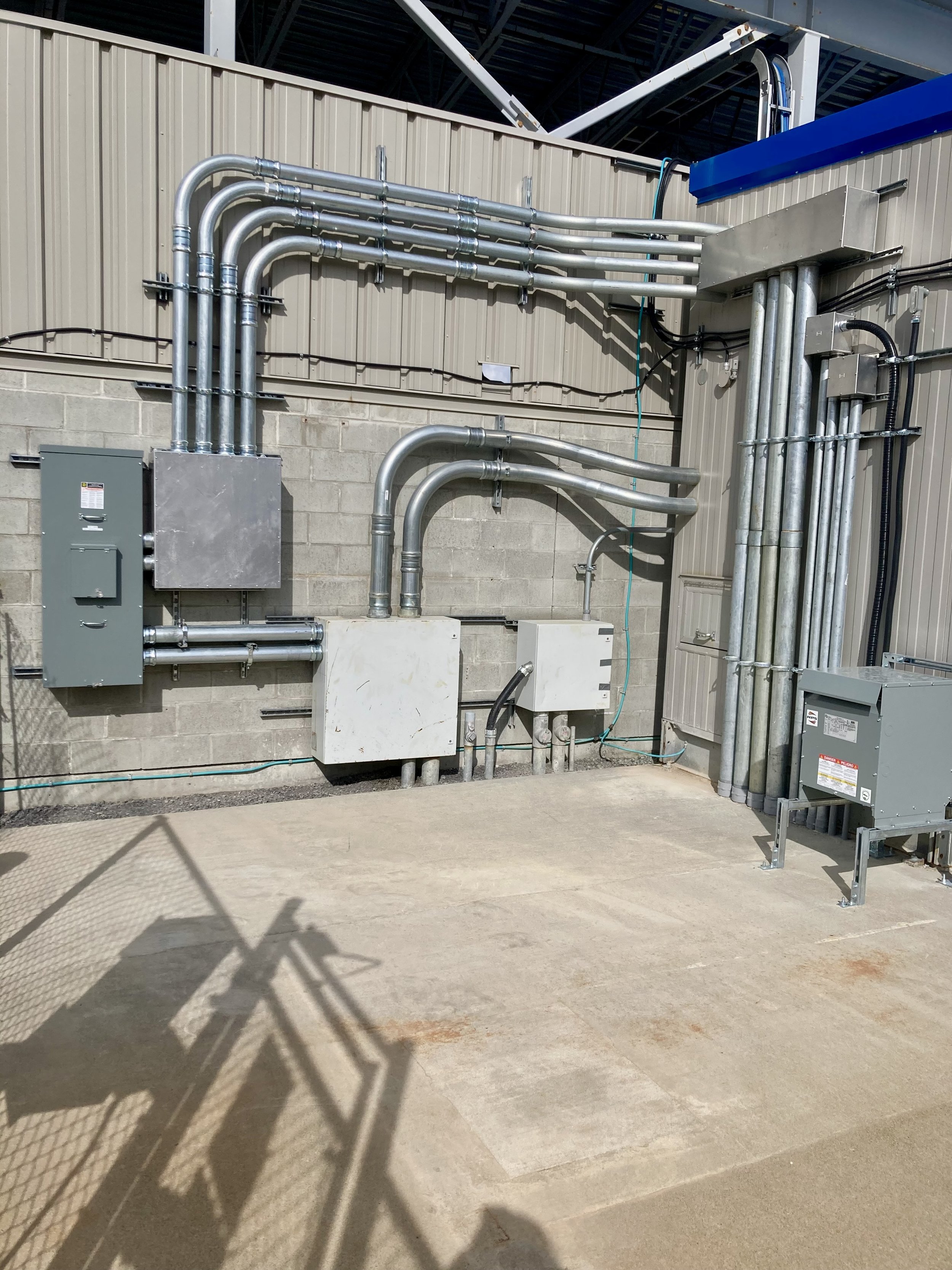 Industrial electrical and piping infrastructure against a concrete wall with electrical cabinets and conduit pipes.