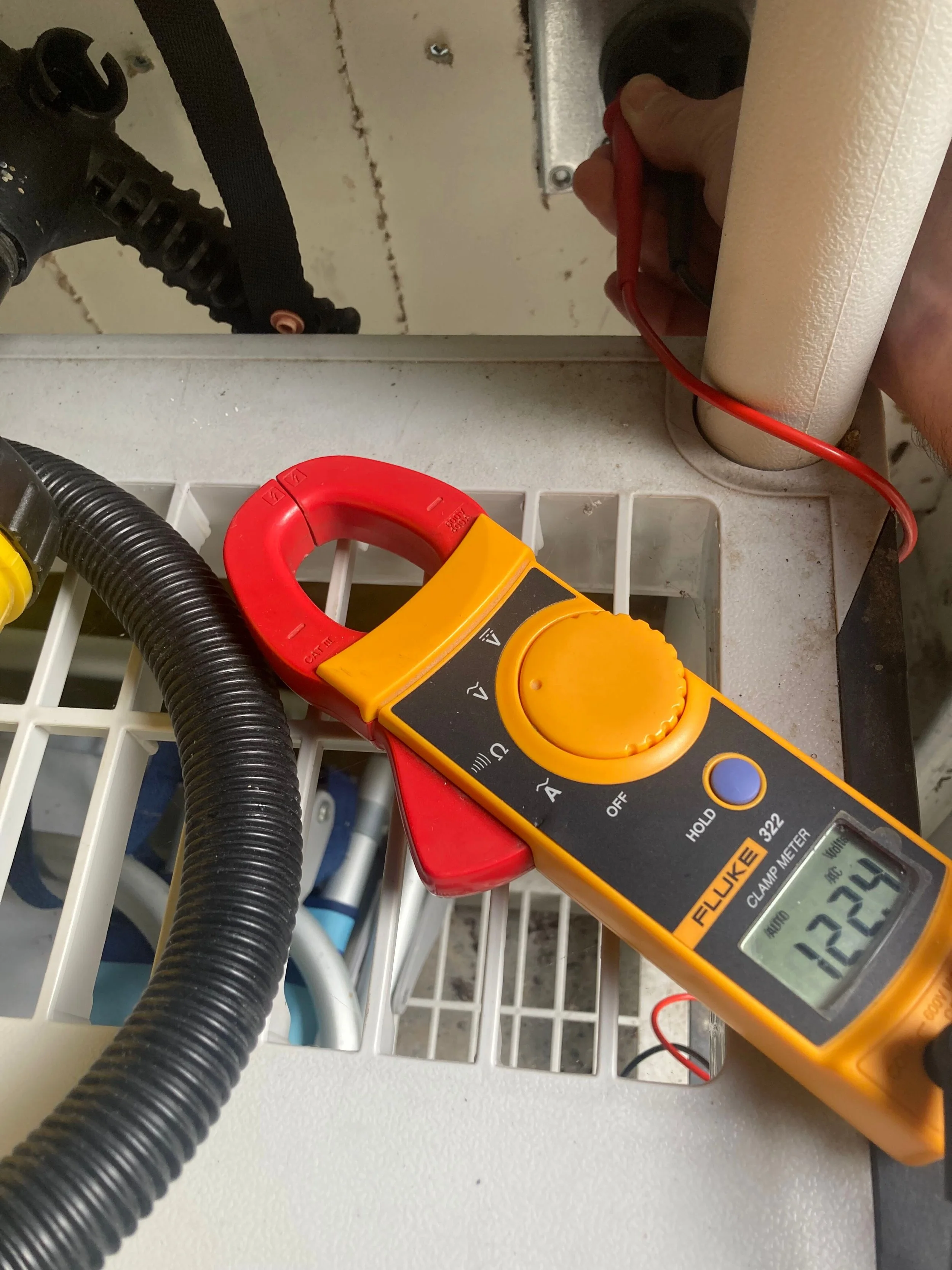 A digital clamp meter measuring 122.4 volts is clamped onto a wire in an electrical panel or device, with a hand holding a red probe near a cylindrical component.