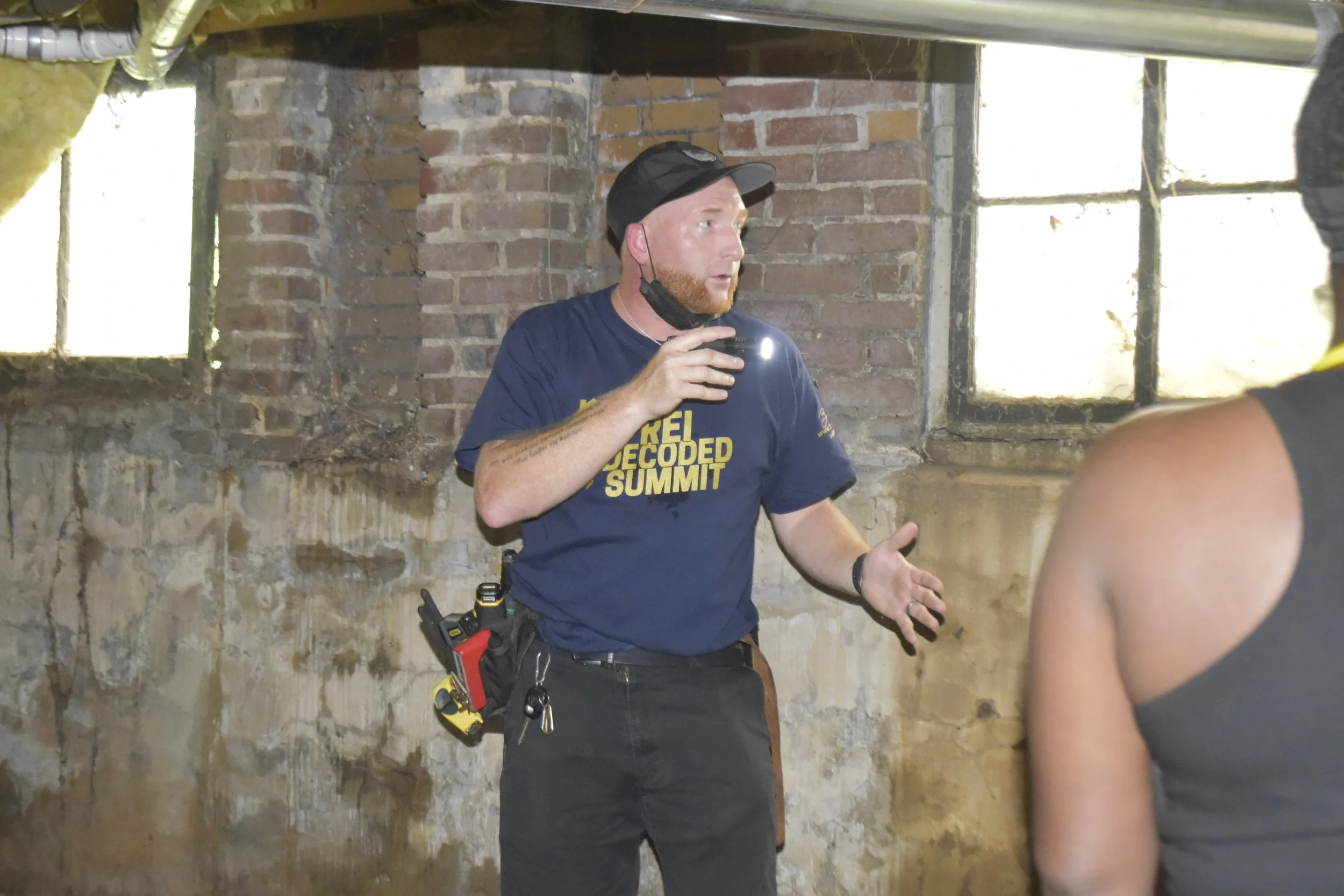 A man in a navy blue t-shirt with the words 'REI Decoded Summit' is speaking or explaining something to a group. He has a black cap, a protective face mask hanging from one ear, and is holding a drink. The man also has a tool belt with various tools attached. The setting appears to be an old brick building with windows, weathered walls, and pouring sunlight.