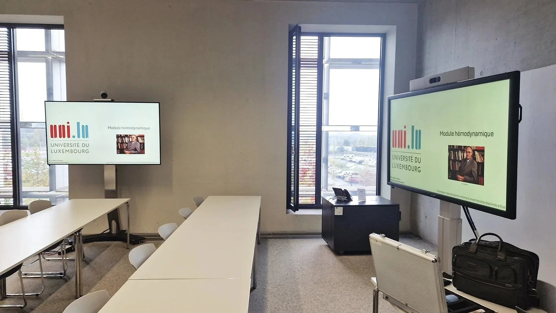 An empty conference room at the University of Luxembourg with two large screens displaying a presentation titled "Module hémodynamique". The room has white tables, white chairs, and large windows letting in natural light.