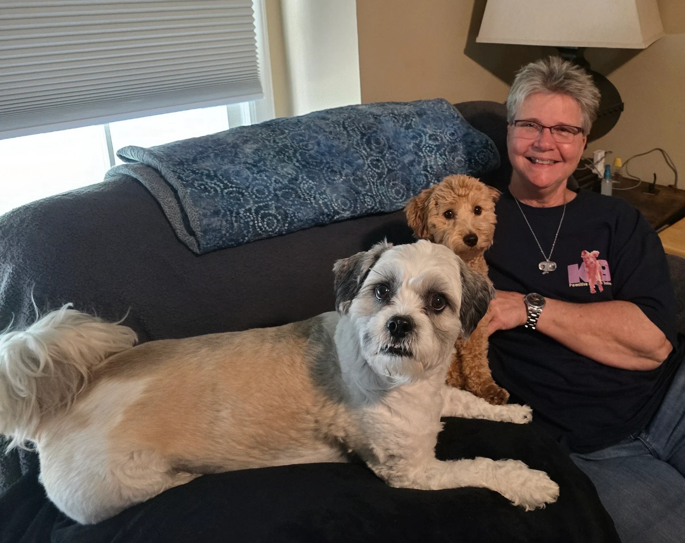 A smiling woman with short gray hair and glasses sitting on a dark gray couch with three dogs in a living room. The woman is wearing a black shirt with pink and purple graphics. The dogs are of various breeds and colors, including a small tan and white dog, a black and white dog with a fluffy coat, and an orange curly-haired dog.