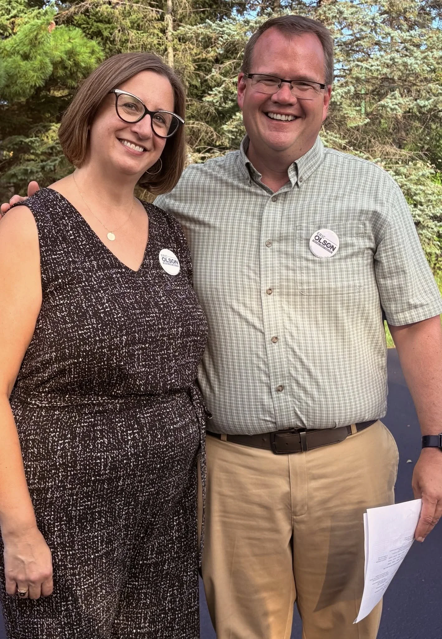 Two people standing outdoors, smiling, wearing campaign stickers that say "Eric Olson", with trees and a blue sky in the background.