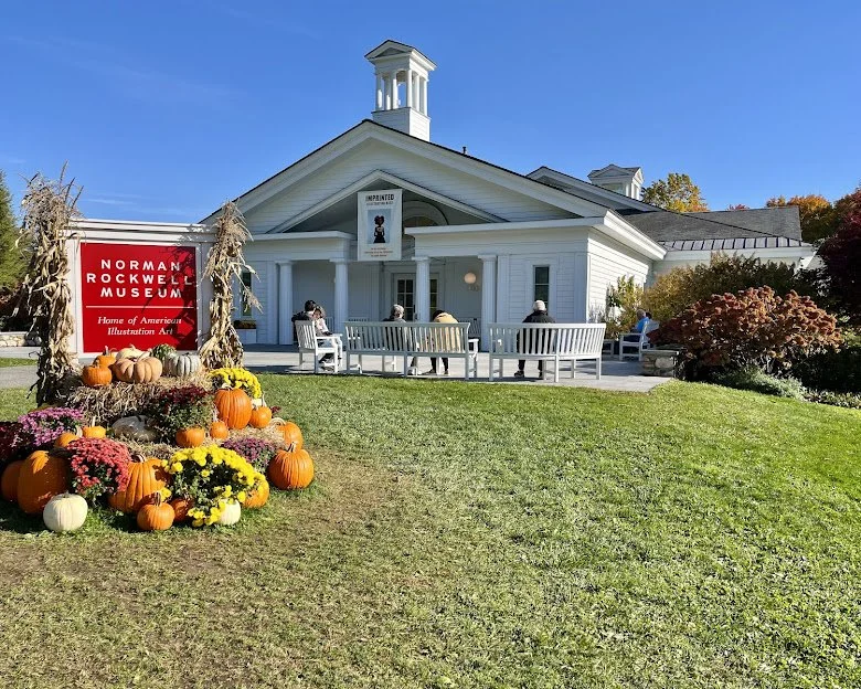 People sitting on benches outside the Norman Rockwell Museum, decorated for fall with pumpkins and mums in front of a white building with a small tower and a sign that says 'Home of Americana Illustration Art'.