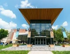 Modern building with a large overhanging roof and glass facade, surrounded by a lawn and trees, under a blue sky.
