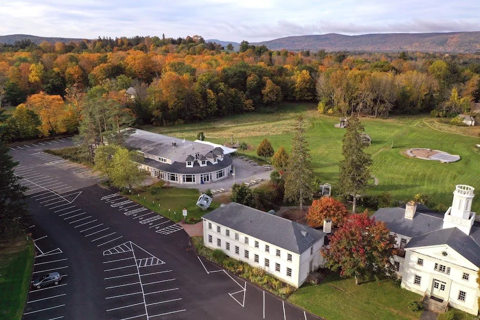 A scenic aerial view of a large estate with a white building, surrounded by a parking lot, lush green fields, a small golf course, and a forested area with colorful autumn trees in the background.