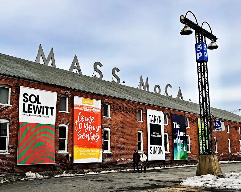 Exterior of a brick building with large banners hanging on the wall, and metal letters spelling 'MASS MoCA' on the roof; sidewalk with two people walking, snow on the ground, and a tall light pole with parking signs.