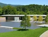 A modern dam and waterfall in a green landscape with trees and hills in the background.