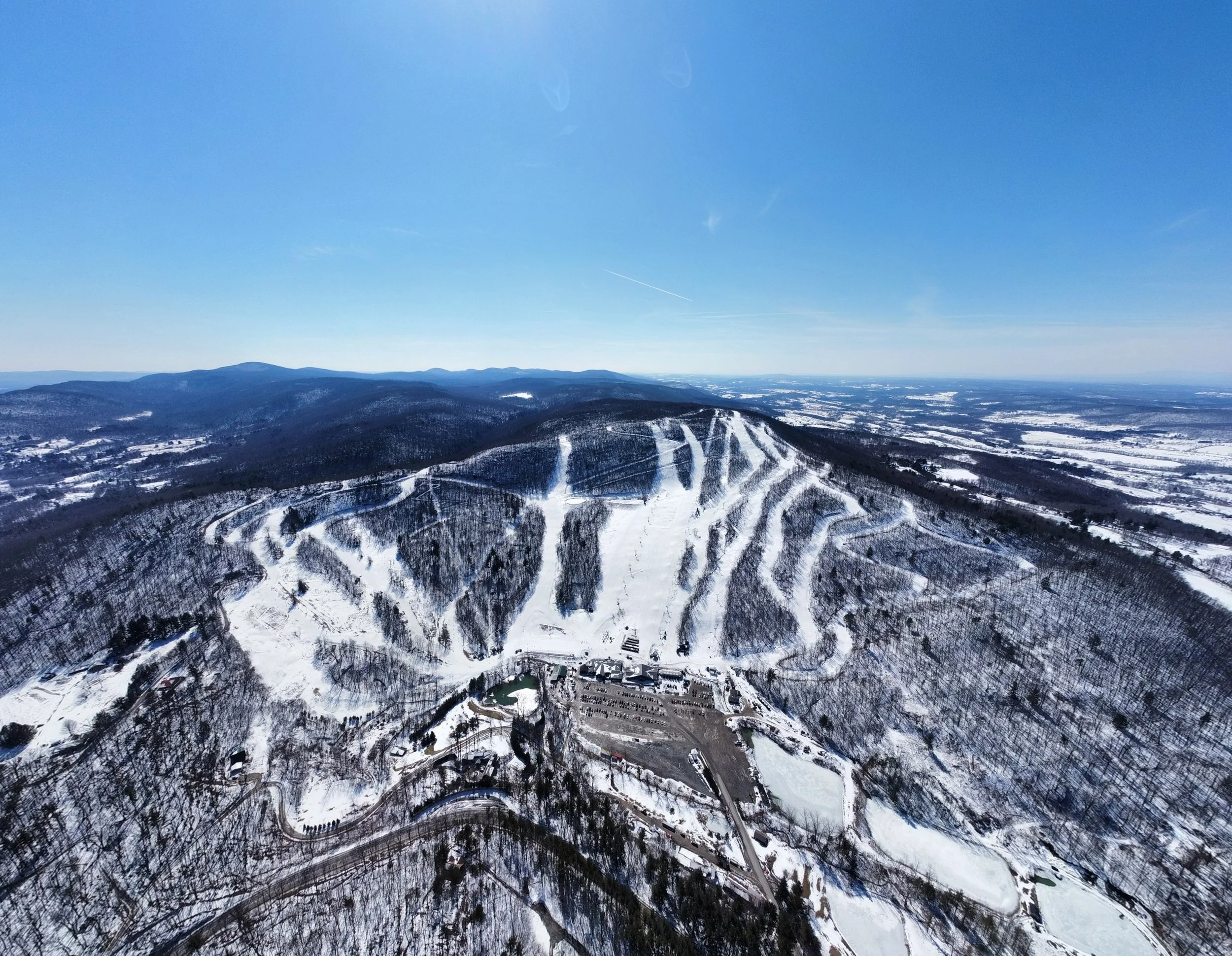 Aerial view of snow-covered ski resort on a mountain, with ski slopes and trails visible under a clear blue sky.