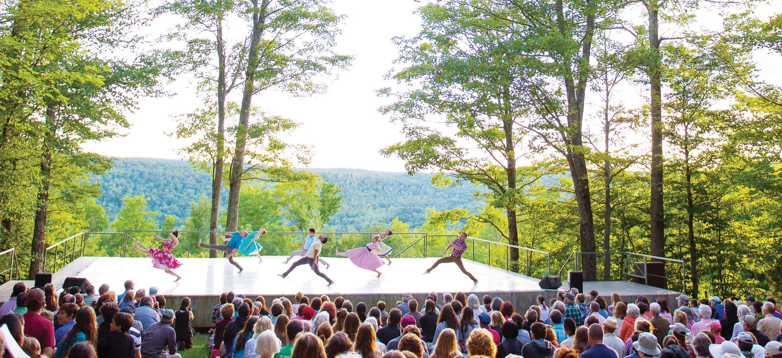 A dance performance on an outdoor stage with a scenic forest and mountain backdrop, audience seated watching