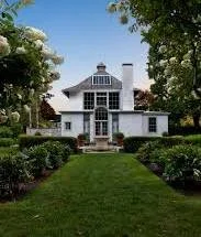 Front view of a large, white house with a gray roof, large windows, and a pathway leading to the entrance, surrounded by a well-maintained lawn and trees.