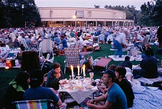People sitting on the grass at Tanglewood, and picnic blankets at an outdoor BSO concert, with tables, candles, and food, in front of a large round stage at dusk.