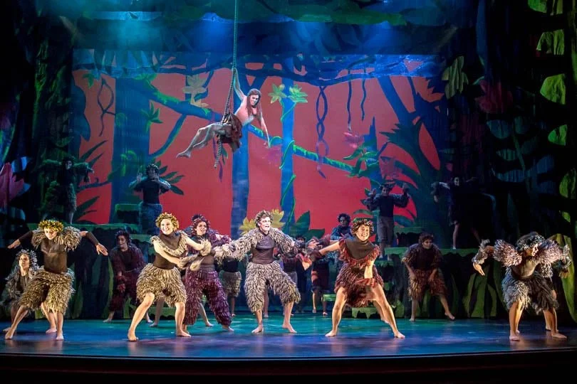 Stage scene from the musical 'The Lion King' featuring performers in animal costumes, some as lions, on a jungle-themed set with a large backdrop of trees and foliage.