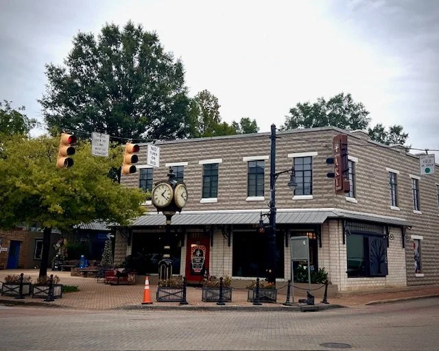 A corner building with a vintage clock, traffic signals, and a small outdoor seating area with benches and trees in a small downtown area.