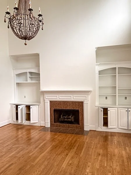 Empty living room with hardwood floors, white built-in shelves, a fireplace with a brown mantel, and a chandelier hanging from the ceiling.
