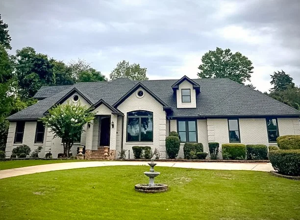 Front view of a large, white, two-story house with a dark roof, landscaping, and a grass lawn with a decorative fountain.