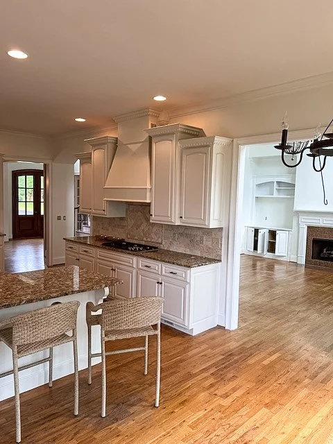 Kitchen with white cabinets, granite countertops, and a gas stove. Adjacent open living room with hardwood floors, a white built-in bookshelf, and a fireplace.