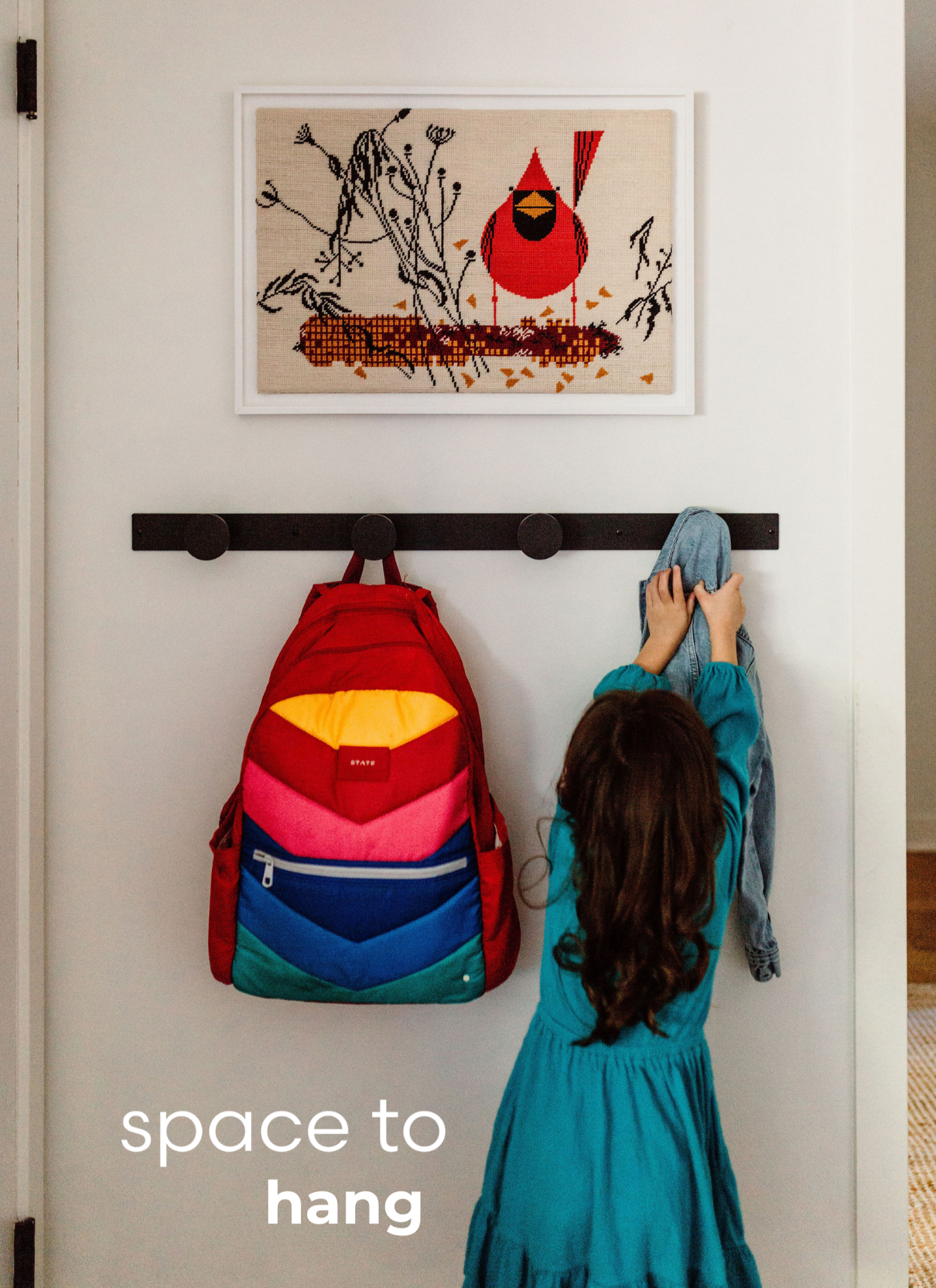 A young girl with long brown hair hanging her jacket and walking toward a wall with hooks, a red backpack hanging on one hook, and a girl reaching for the jacket on another hook, with a white wall and a framed bird-themed artwork above.