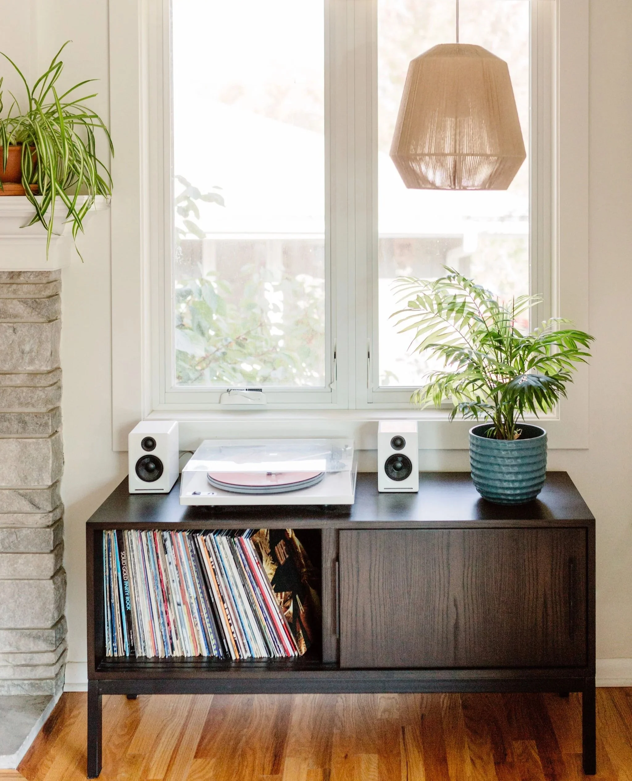 A modern living room with a black console table containing vinyl records, a white record player, and two white speakers. The table is placed beneath a window with a view of green foliage outside. On the left side, there is a plant on a shelf above a 