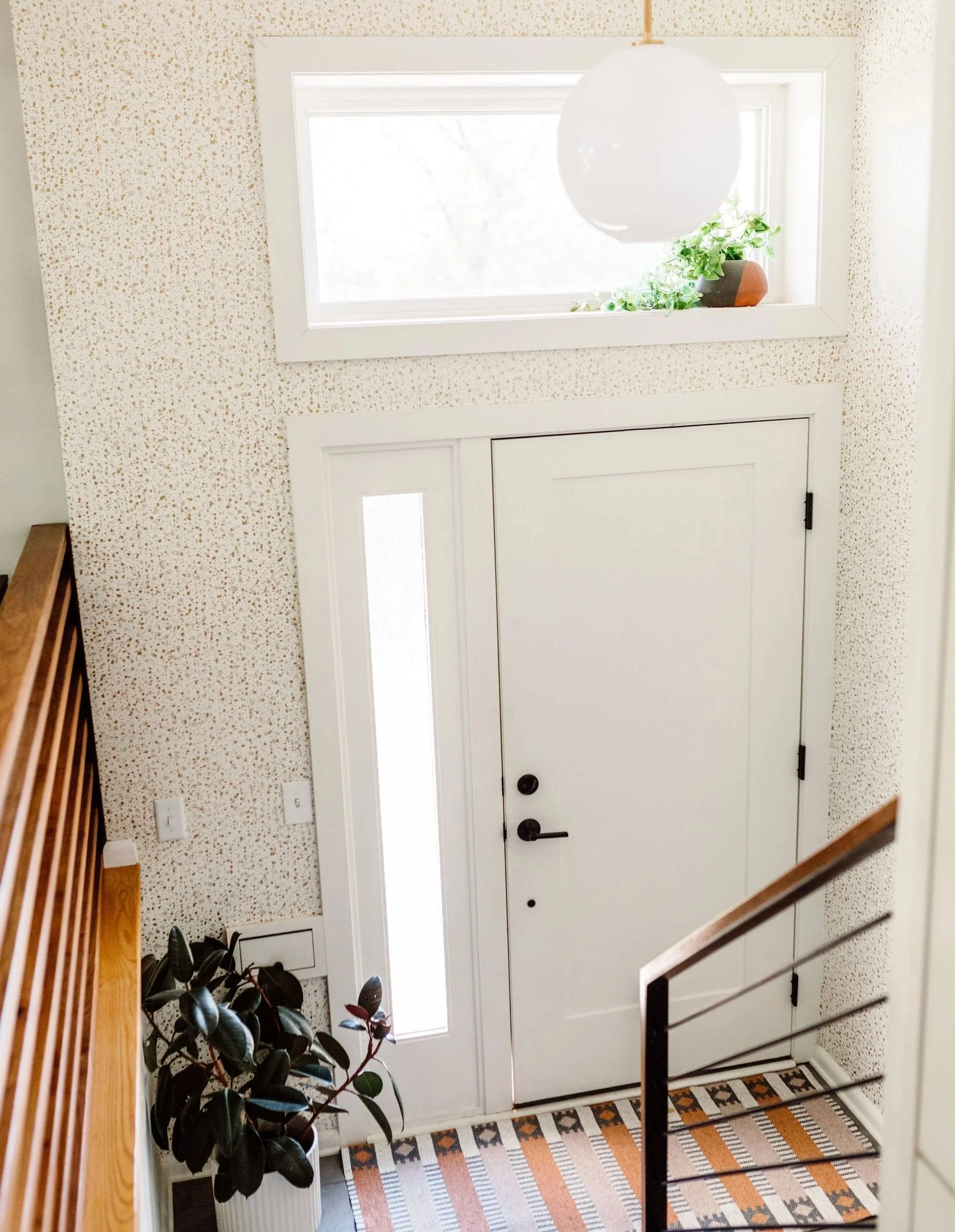 Entryway with white door, sidelights, hanging globe pendant light, potted plant, patterned rug, and staircase railing.