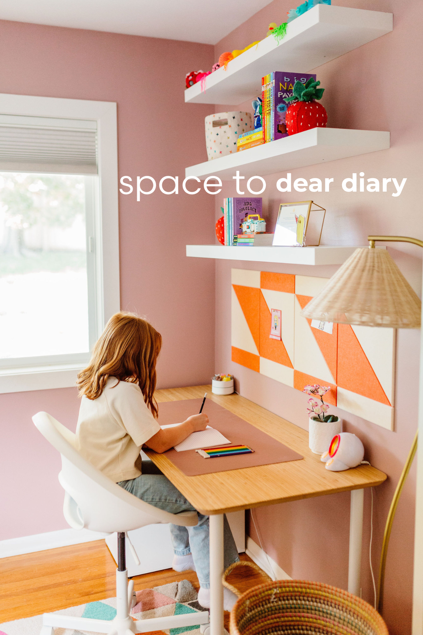 A young girl with red hair sitting at a wooden desk, writing in a notebook. The wall behind her is pink with two white shelves holding colorful toys and books, and an orange and white geometric art piece. There is a window to her left with a view out