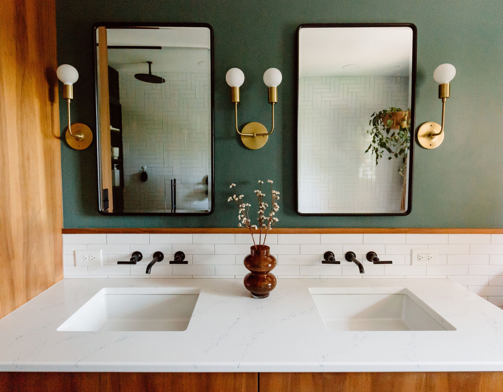 A bathroom double vanity with white countertop, two square undermount sinks, two black wall-mounted faucets, two large rectangular mirrors, two gold wall sconces with white globes, a brown vase with dried flowers, a green wall background, and white t