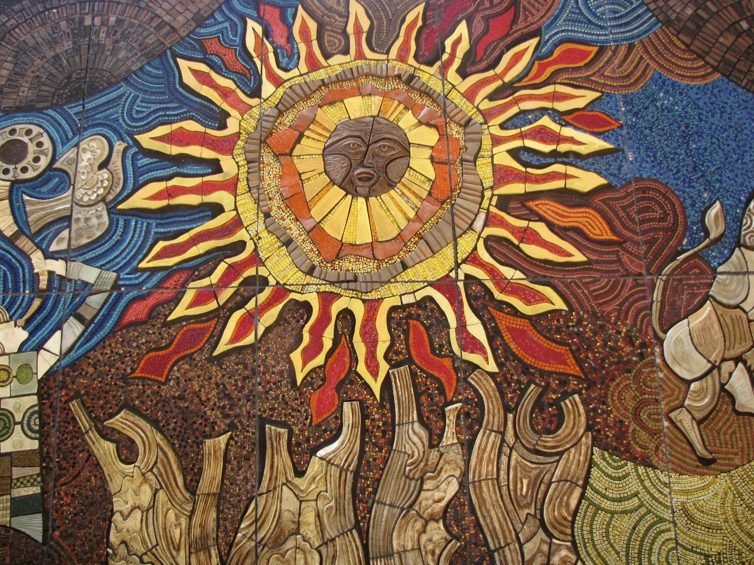 A vibrant mosaic mural featuring a central sun with a human face, surrounded by colorful flames, blue and red patterns, and abstract designs.