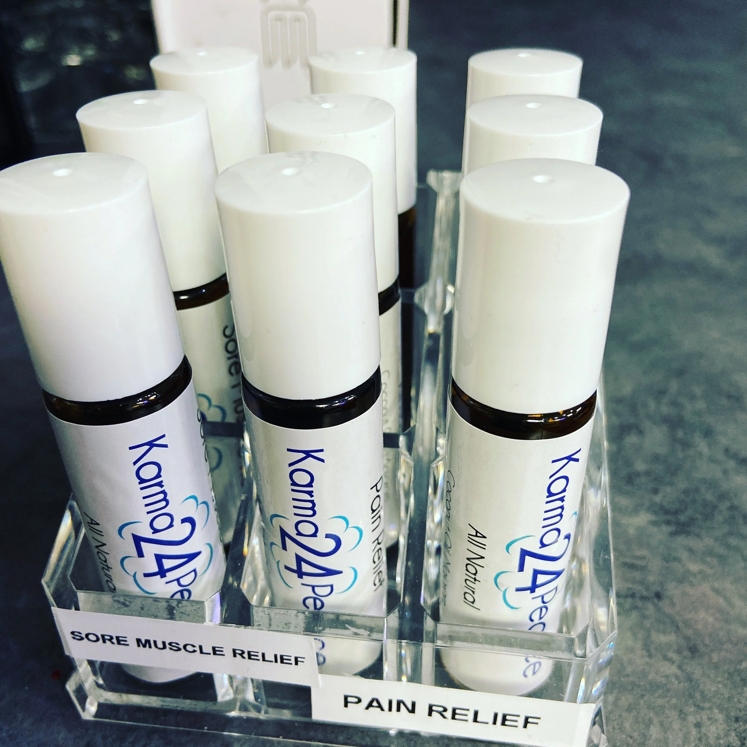 Lip balm tubes labeled with 'Karma24 Peace 'All Natural', and displayed in a clear acrylic holder with signs for 'Sore Muscle Relief' and 'Pain Relief'.