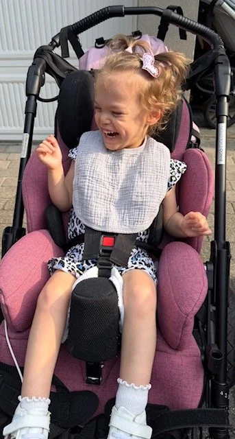 A young girl with curly hair in pigtails, wearing a gray bib and a patterned dress, sitting in a pink stroller and laughing cheerfully.