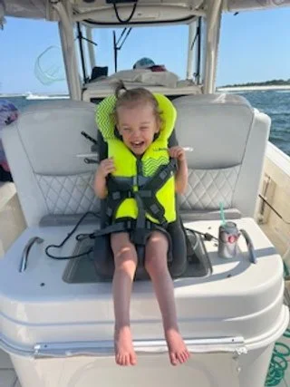 Child smiling and enjoying a boat ride while wearing a bright yellow life jacket, sitting on the back of a boat with water and sky in the background.