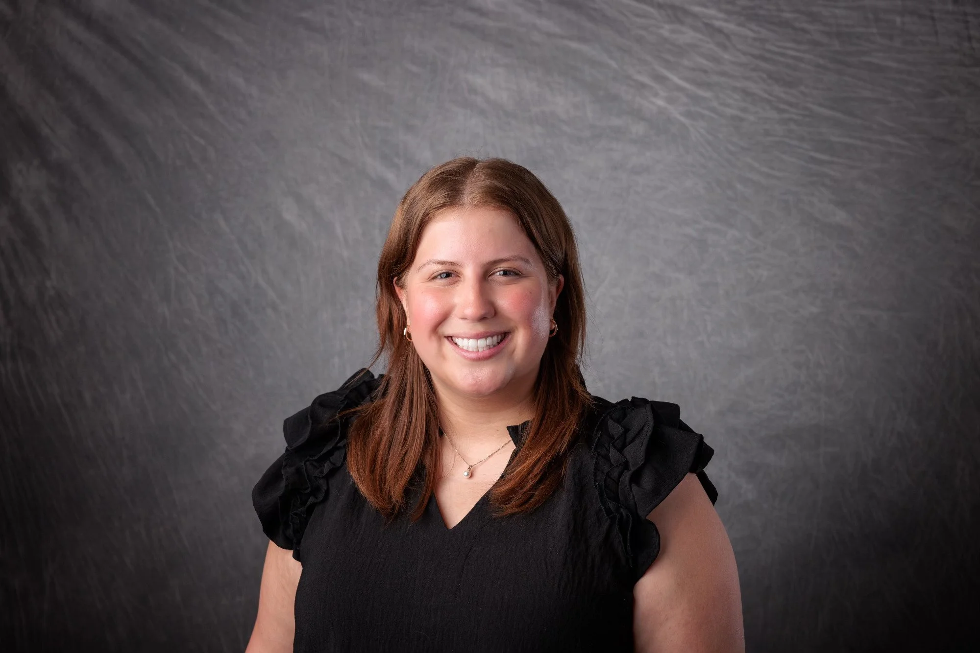 A young woman with shoulder-length brown hair smiling, wearing a black top with ruffled sleeves, a necklace, and earrings, standing against a textured gray background.