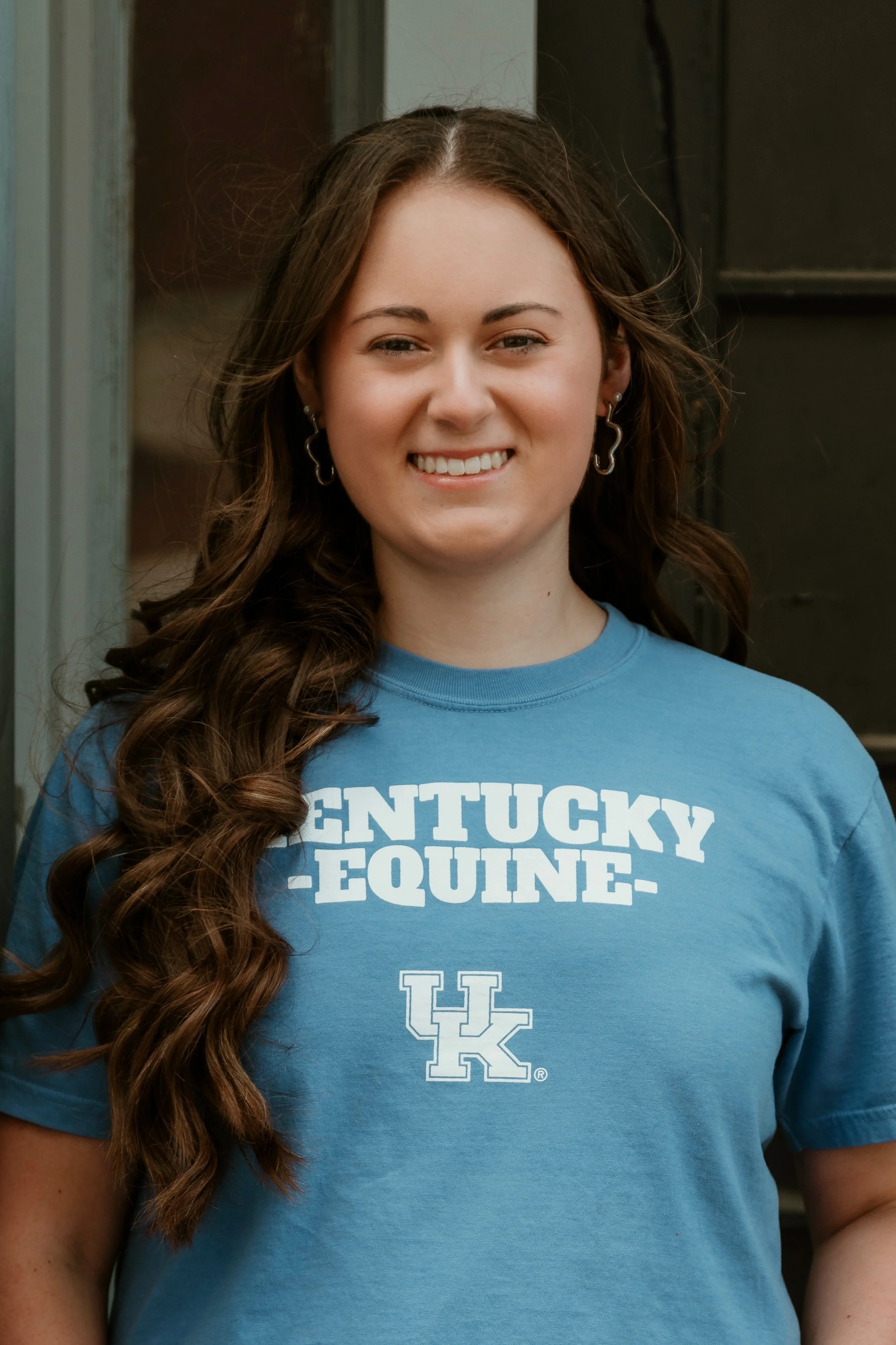 A young woman with long, wavy brown hair wearing a blue Kentucky Equine T-shirt and hoop earrings, smiling at the camera.