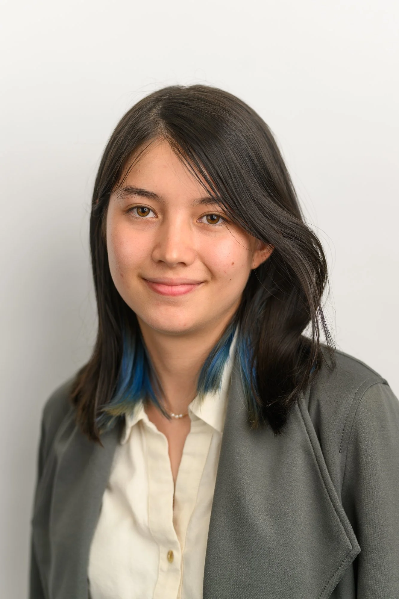 A young woman with dark hair and a small streak of blue, smiling softly at the camera, wearing a grey blazer over a white shirt, with a simple necklace, standing against a plain white background.