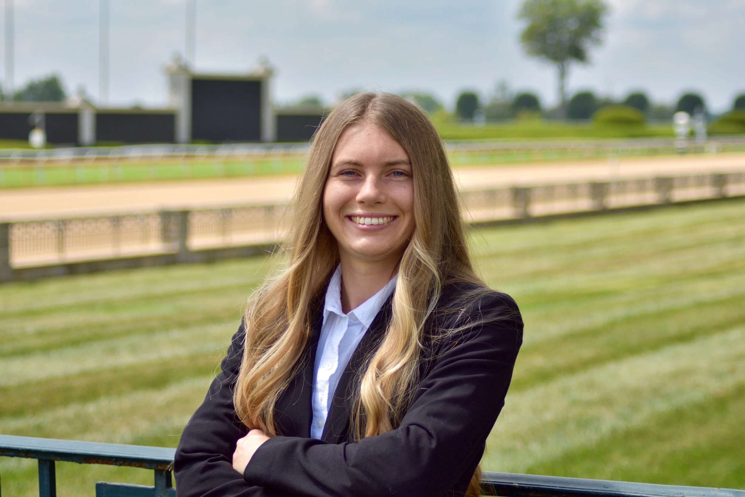 Young woman with long blonde hair wearing a black blazer and white shirt, standing in front of a grassy race track with trees and fences in the background.